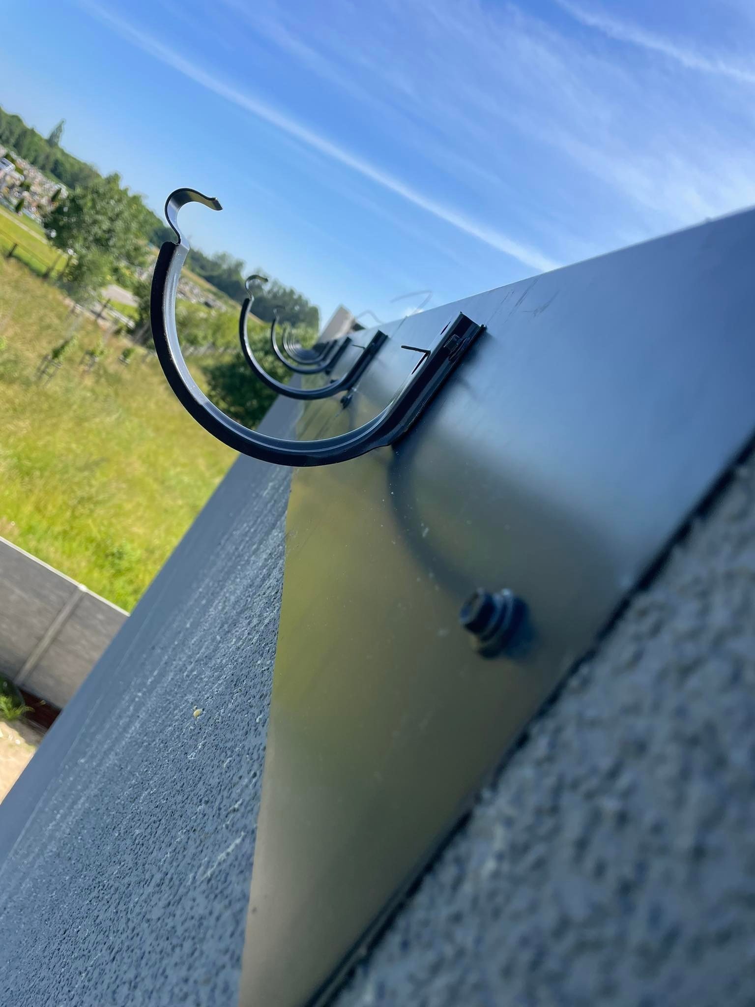 Czarne, metalowe uchwyty rynnowe zamocowane do ciemnej, gładkiej powierzchni dachu, widok z dołu na błękitne niebo i zieleń w tle.