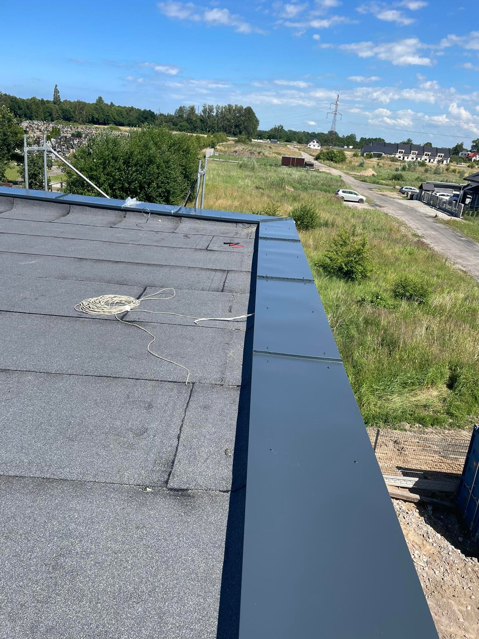 Widok z góry na fragment dachu pokrytego papą, z zamontowanymi, szarymi obróbkami blacharskimi przy krawędzi, zwinięty kabel i narzędzia na powierzchni, w tle krajobraz z drzewami i domami.