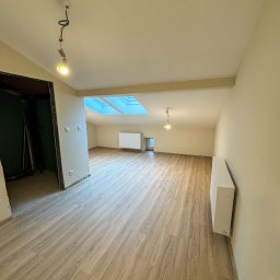 SIMPLE FLAT SPÓŁKA Z OGRANICZONĄ ODPOWIEDZIALNOŚCIĄ - Wnętrze pokoju po remoncie w Nowym Sączu z panelami podłogowymi w odcieniach drewna, widoczne grzejniki, okno dachowe i wisząca żarówka bez klosza.