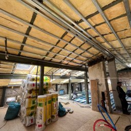 SIMPLE FLAT SPÓŁKA Z OGRANICZONĄ ODPOWIEDZIALNOŚCIĄ - Montaż izolacji termicznej z wełny mineralnej Isover w poddaszu, widoczne metalowe profile sufitowe, stos rolek izolacji, oraz pracownicy podczas instalacji.