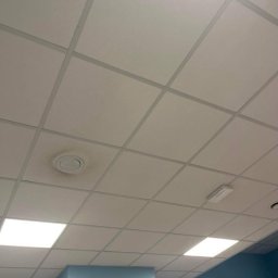 SIMPLE FLAT SPÓŁKA Z OGRANICZONĄ ODPOWIEDZIALNOŚCIĄ - Kasetonowy sufit podwieszany z widocznym zabrudzeniem wokół kratki wentylacyjnej oraz fragment niebieskiej ściany.