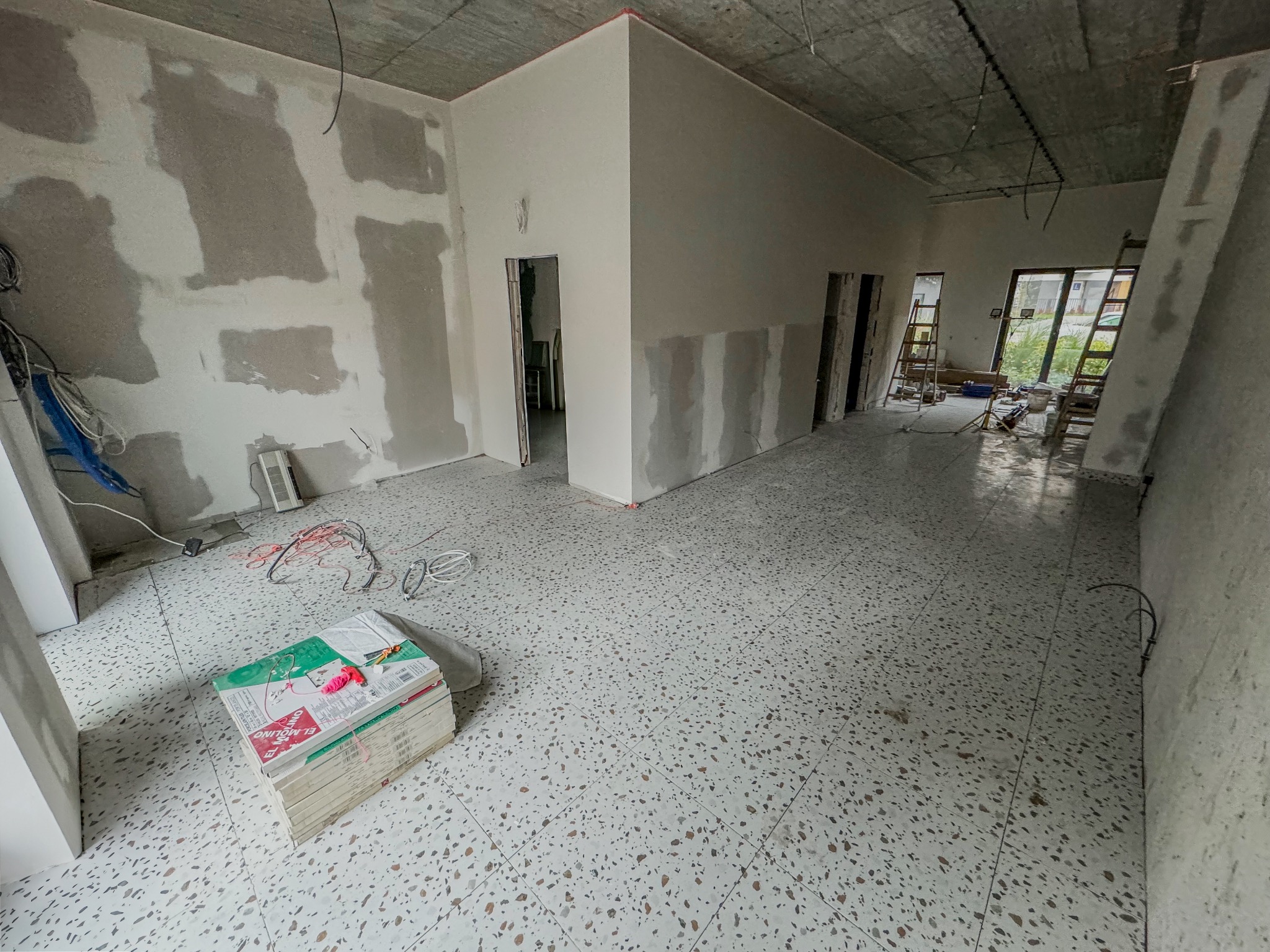 Wnętrze w trakcie remontu: ściany częściowo wygładzone gładzią, widoczne kable, podłoga z lastryko, drzwi bez ościeżnic, stos płytek podłogowych na podłodze.