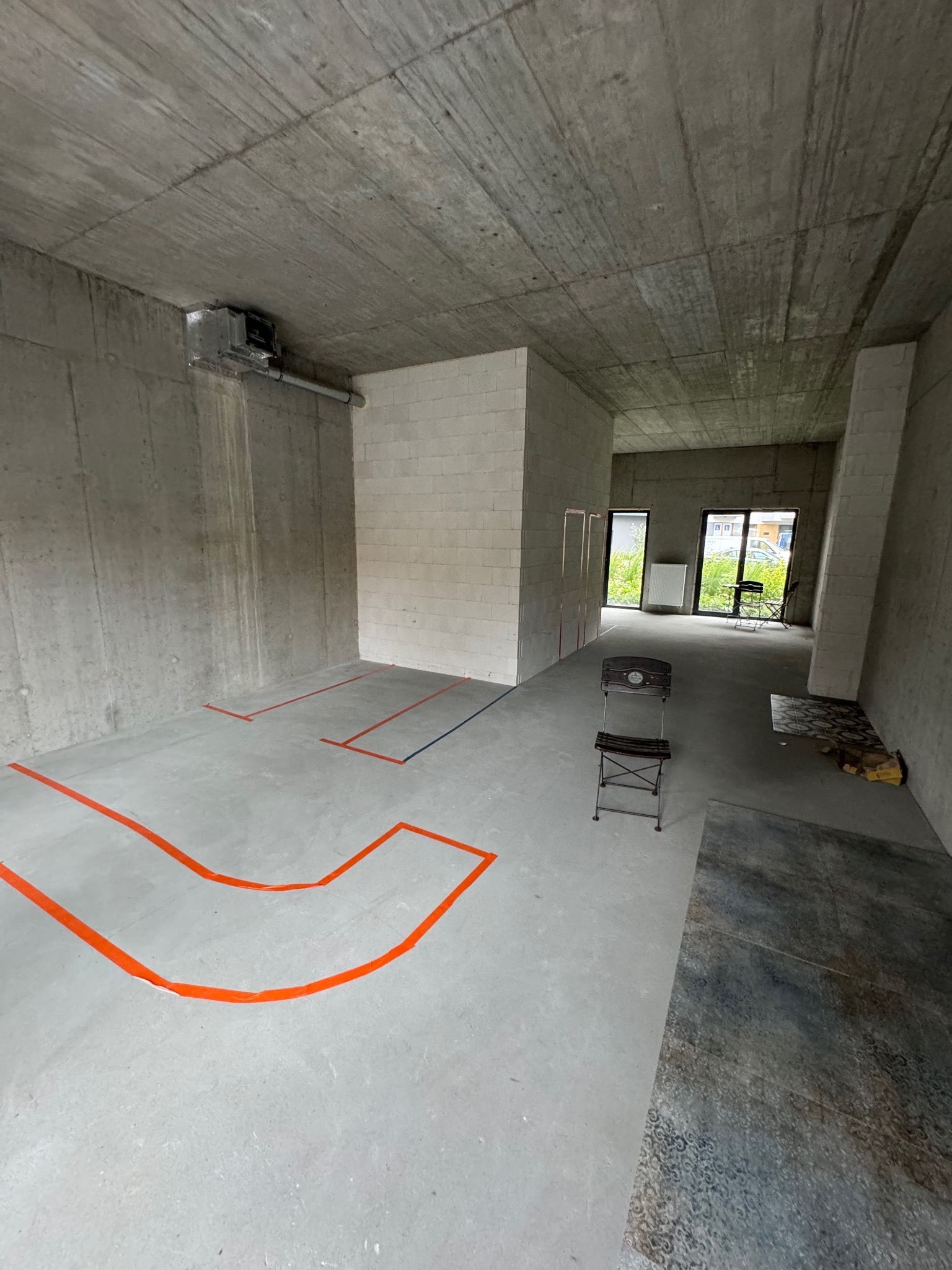 Surowy stan wnętrza: betonowe ściany i strop, zaznaczone pomarańczową taśmą linie na podłodze, fragment ułożonych płytek podłogowych, krzesło w tle. Widoczne okna z zielenią na zewnątrz.