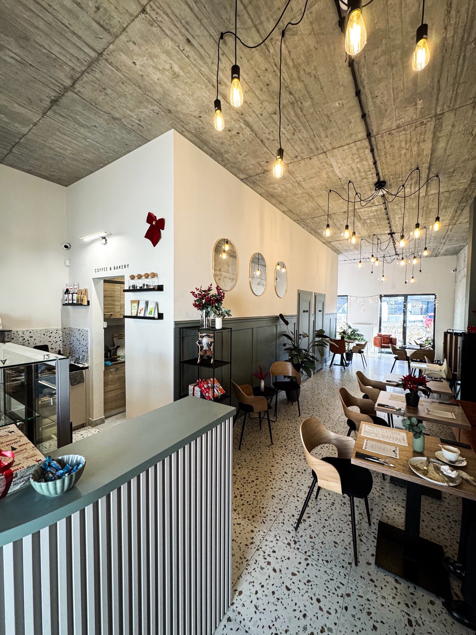 Wnętrze kawiarni z betonowym sufitem, wiszącymi żarówkami, stolikami z drewnianymi krzesłami i podłogą z lastryko. W tle widoczny bar z napisem 'Coffee & Bakery' oraz dekoracje świąteczne.