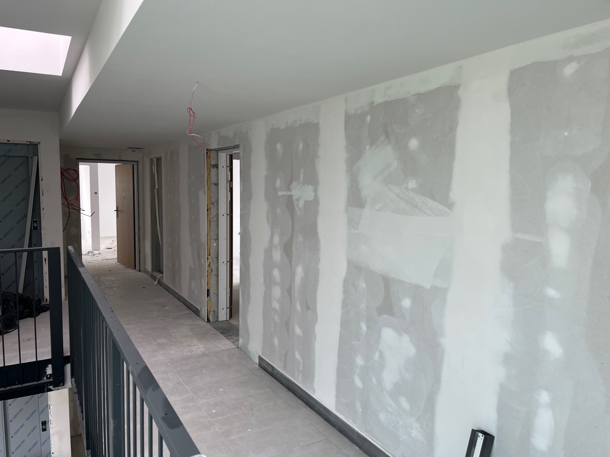 Korytarz w trakcie prac wykończeniowych: ściany z nałożonym gipsem i widocznymi śladami szpachlowania, szare płytki podłogowe, czarna balustrada i widoczne instalacje elektryczne.