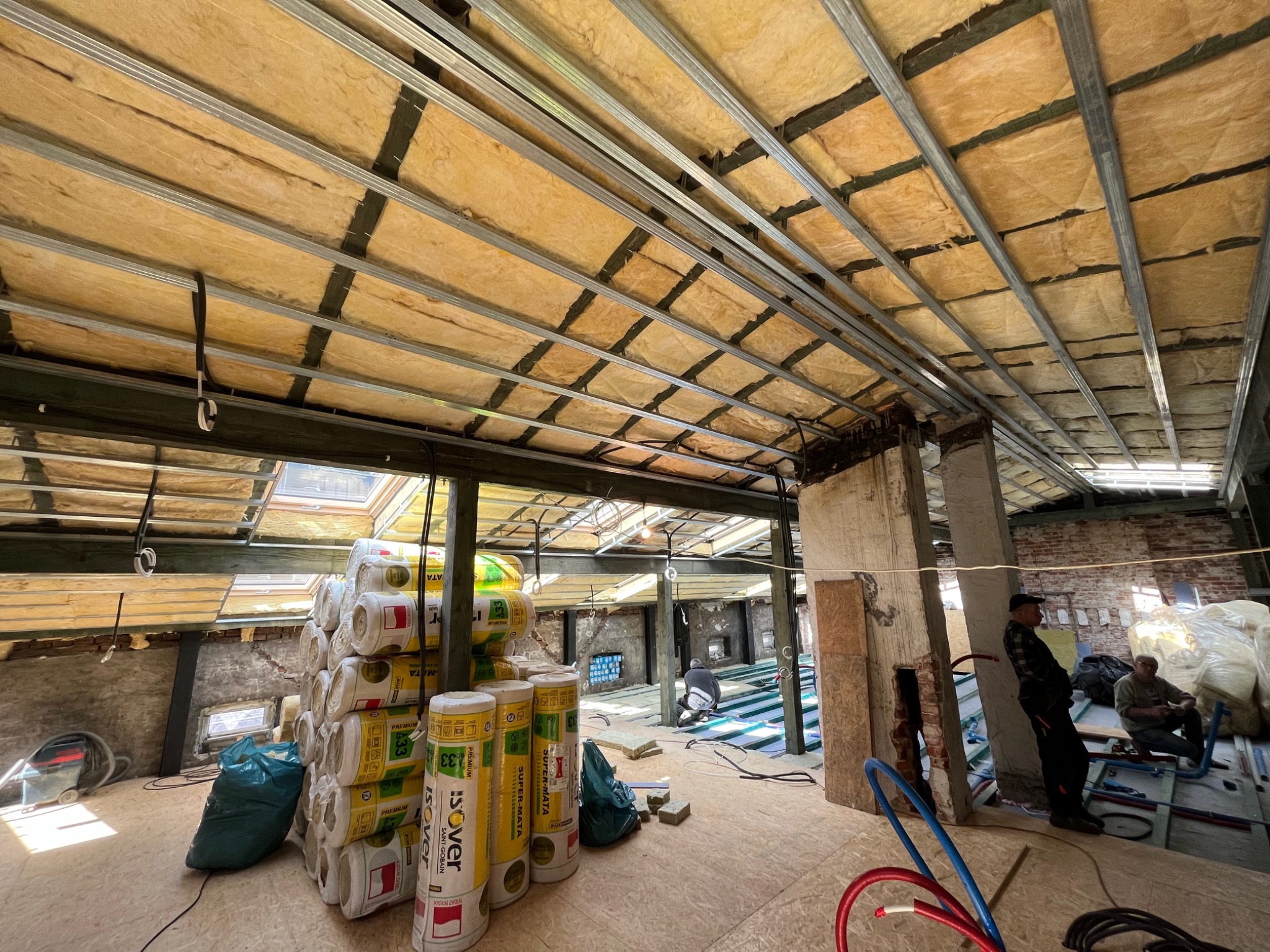 Montaż izolacji termicznej z wełny mineralnej Isover w poddaszu, widoczne metalowe profile sufitowe, stos rolek izolacji, oraz pracownicy podczas instalacji.