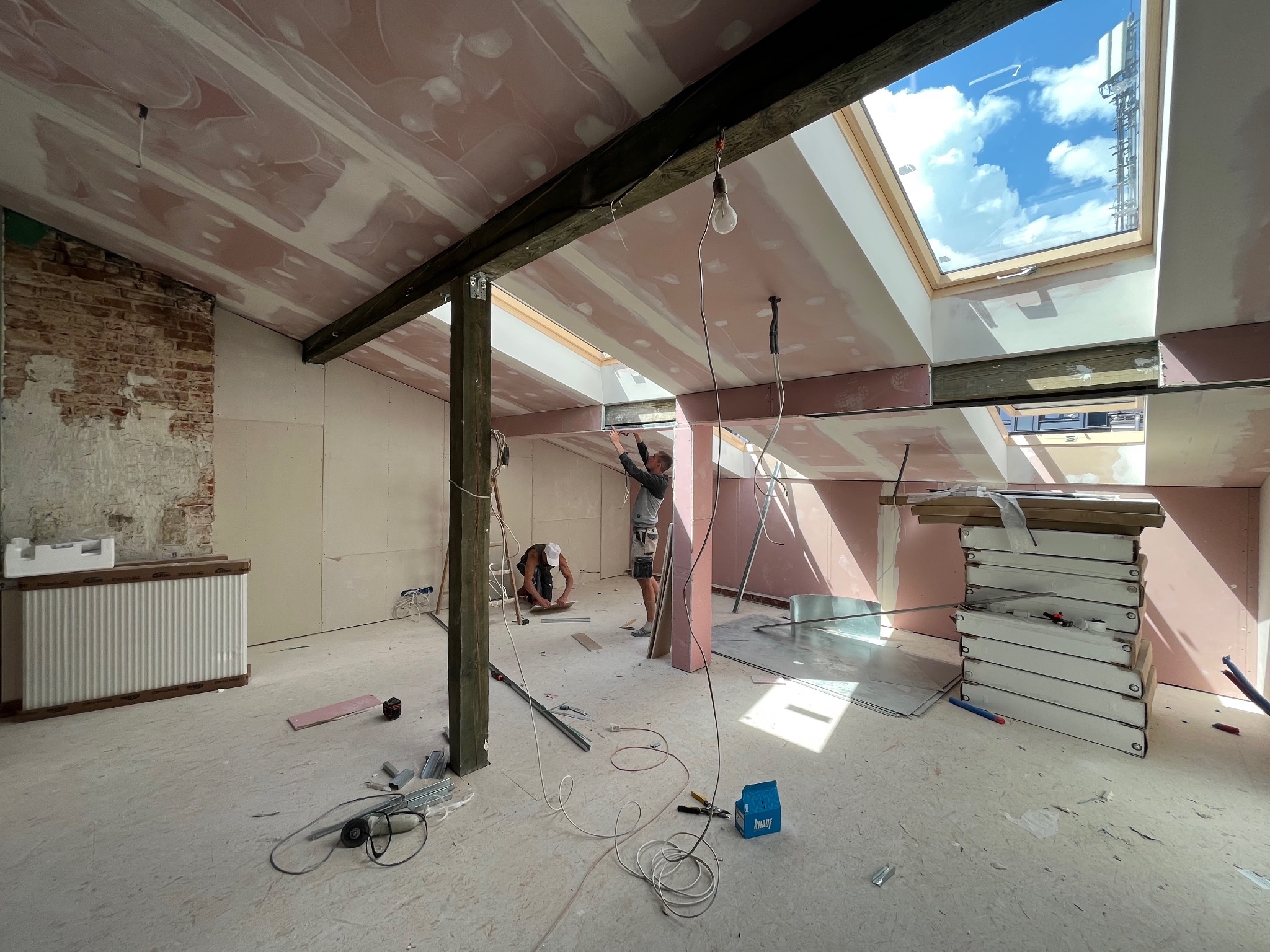 Poddasze w trakcie wykończenia: montaż płyt gipsowo-kartonowych, widoczne drewniane belki stropowe, okno dachowe z widokiem na błękitne niebo, grzejnik w opakowaniu i narzędzia pracy na podłodze.