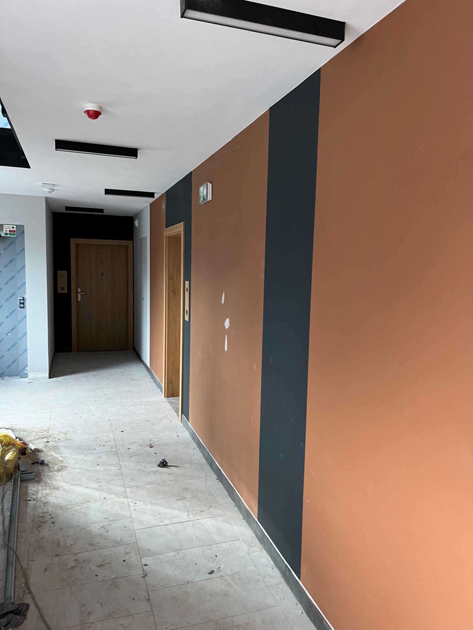 Korytarz w trakcie wykańczania w Nowym Sączu: pomalowane ściany w odcieniach brązu i granatu, widoczne ślady po pracach na podłodze, drzwi do windy i pomieszczeń.