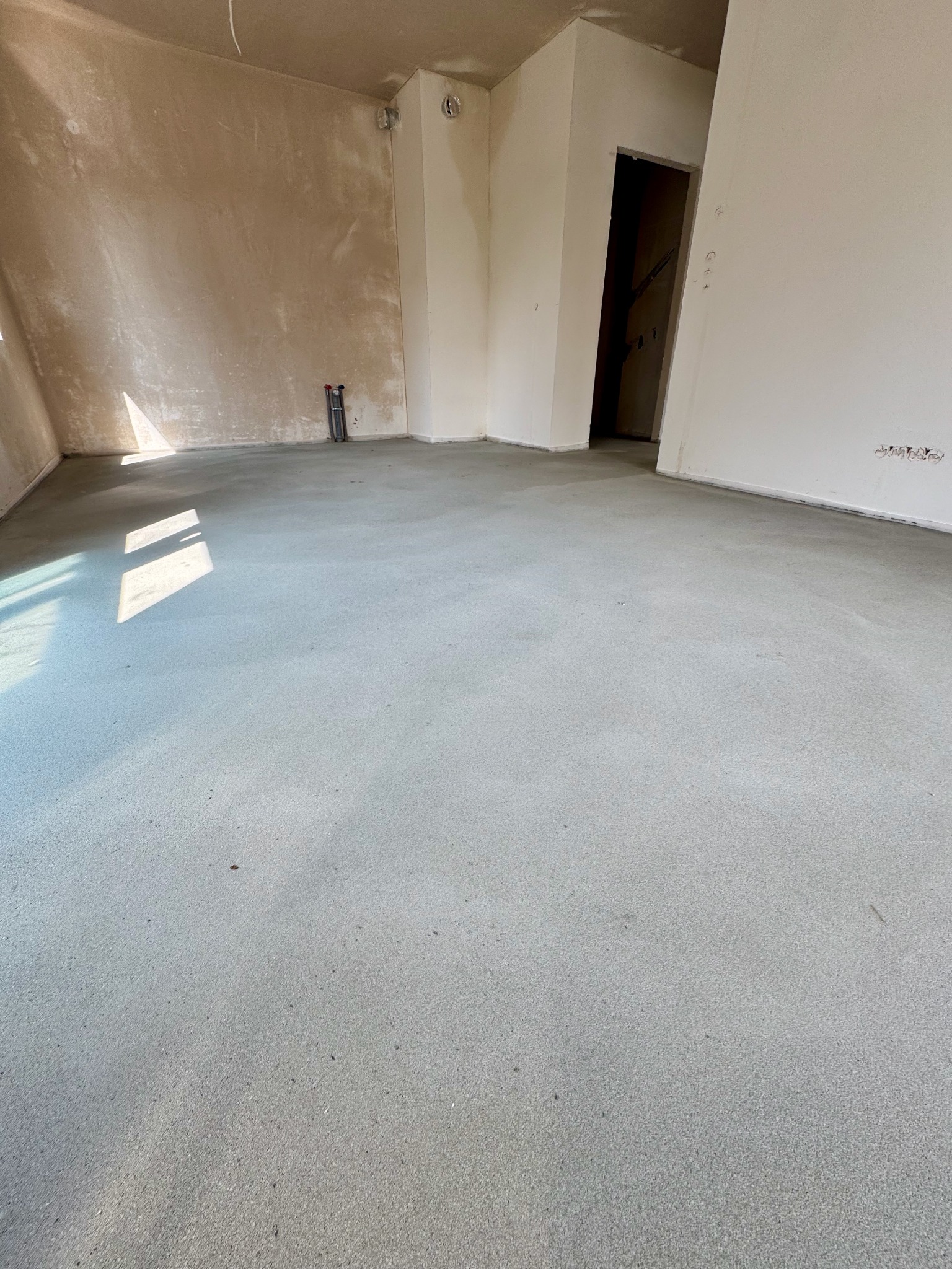 Świeżo wylana, gładka wylewka cementowa w jasnym pomieszczeniu, z widocznymi śladami po narzędziach i rurami w tle. Naturalne światło podkreśla strukturę podłoża.