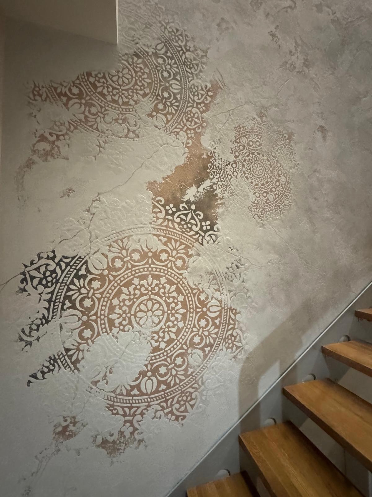 Dekoracyjna ściana z wzorem mandali w odcieniach bieli i brązu, wykonana techniką stiuku, sąsiadująca z drewnianymi schodami z metalową barierką.