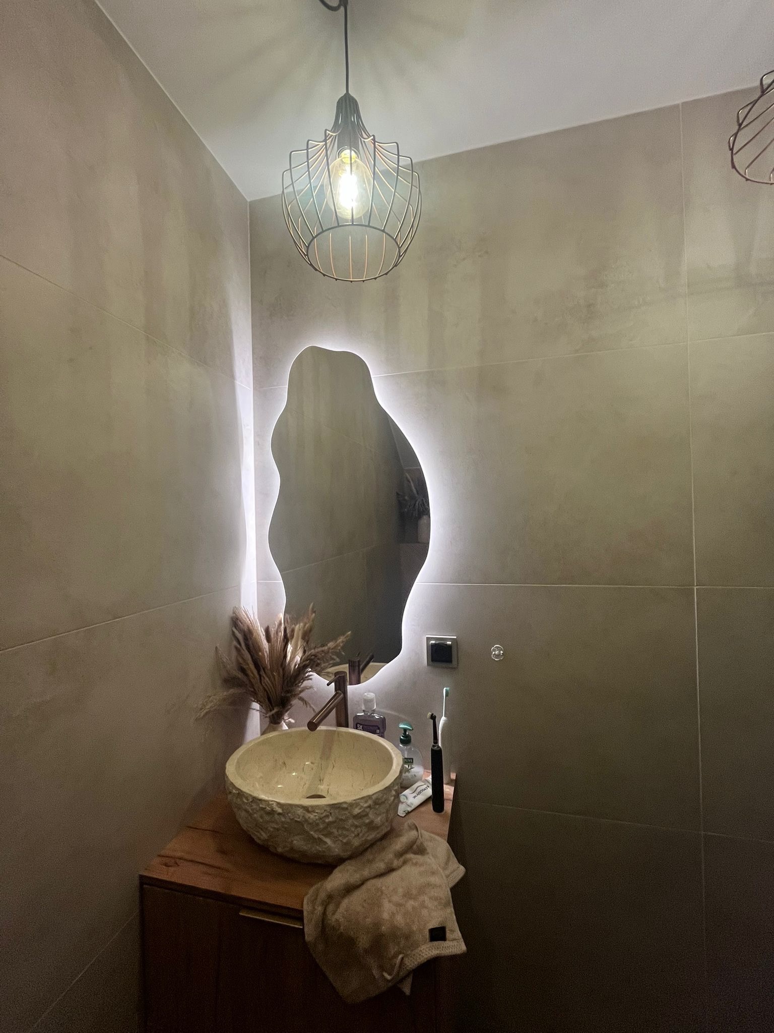 Stylowa łazienka z umywalką z kamienia, podświetlanym lustrem o nieregularnym kształcie i nowoczesną lampą wiszącą. Beżowe płytki na ścianach, drewniana szafka pod umywalką.