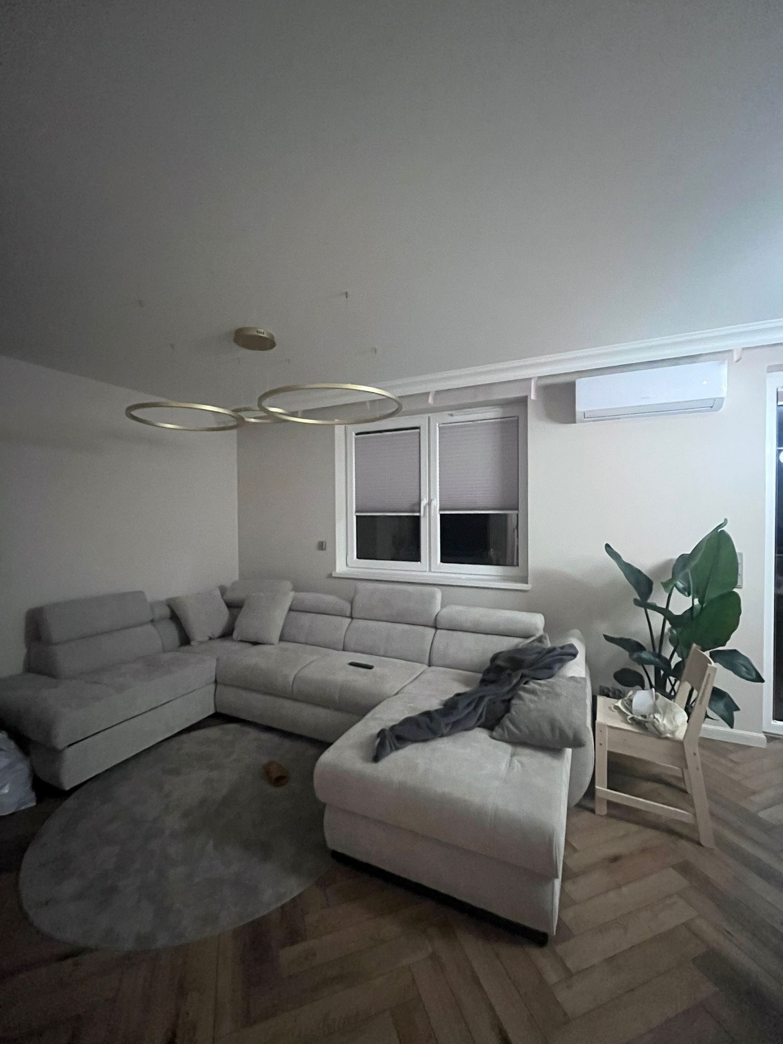 Stylowy salon z narożnikiem w odcieniach szarości, okrągłym dywanem i drewnianą podłogą w jodełkę. Uwagę zwraca nowoczesna lampa sufitowa i klimatyzator.