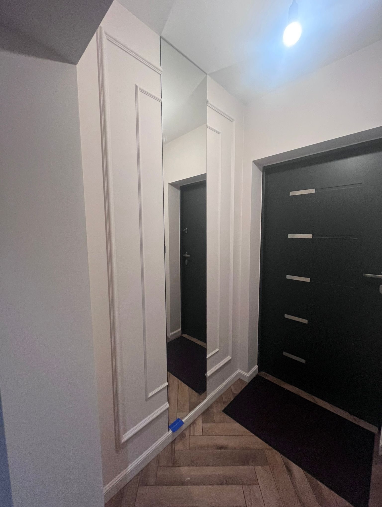 Korytarz po remoncie: lustro w ozdobnej ramie, czarne drzwi z poziomymi paskami, parkiet w jodełkę. Nowoczesne wykończenie wnętrza w jasnych barwach.