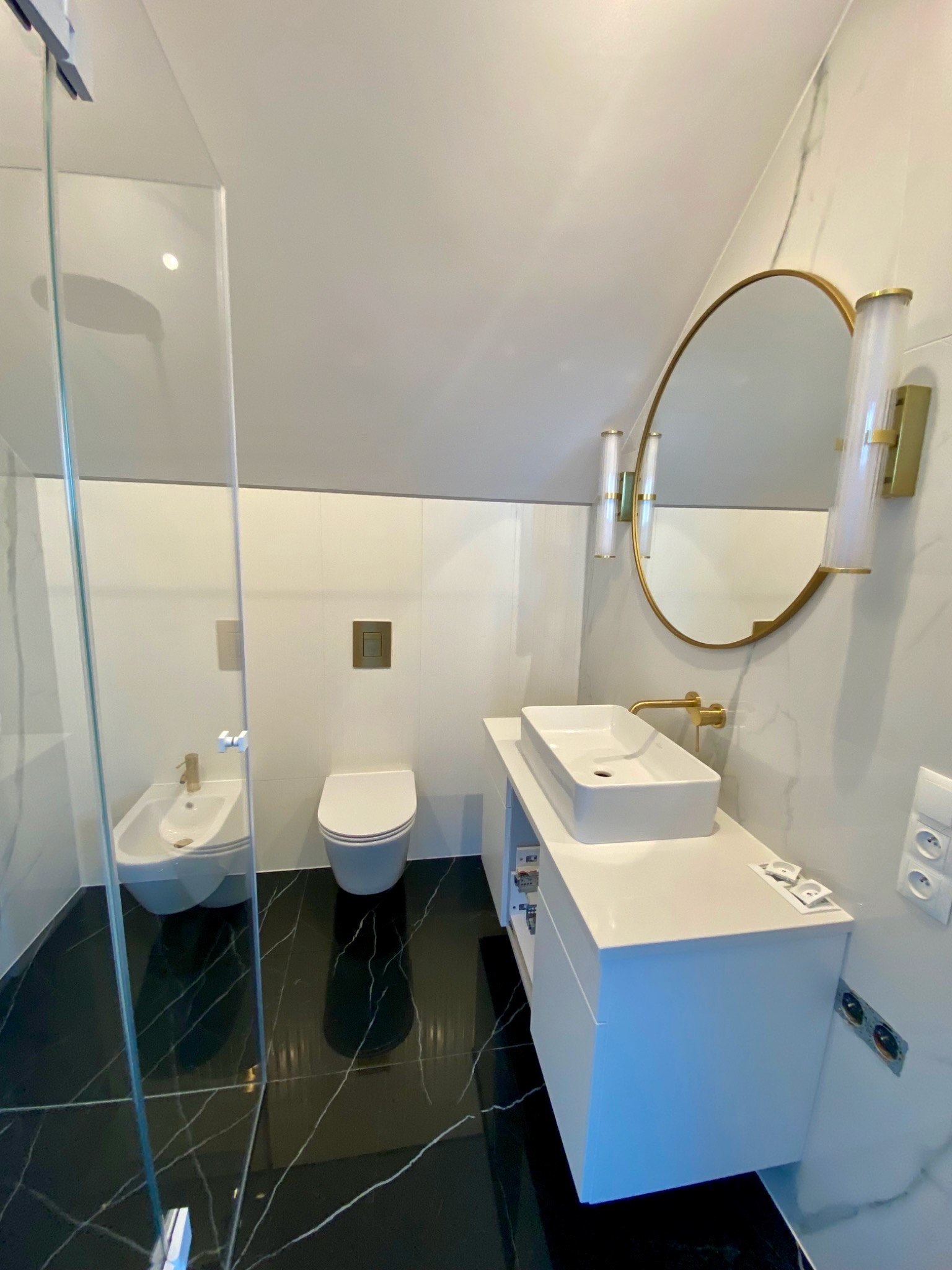 Nowoczesna łazienka z czarną podłogą, białą armaturą i złotymi akcentami: owalne lustro, bateria umywalkowa, oświetlenie. Kabina prysznicowa ze szkła.