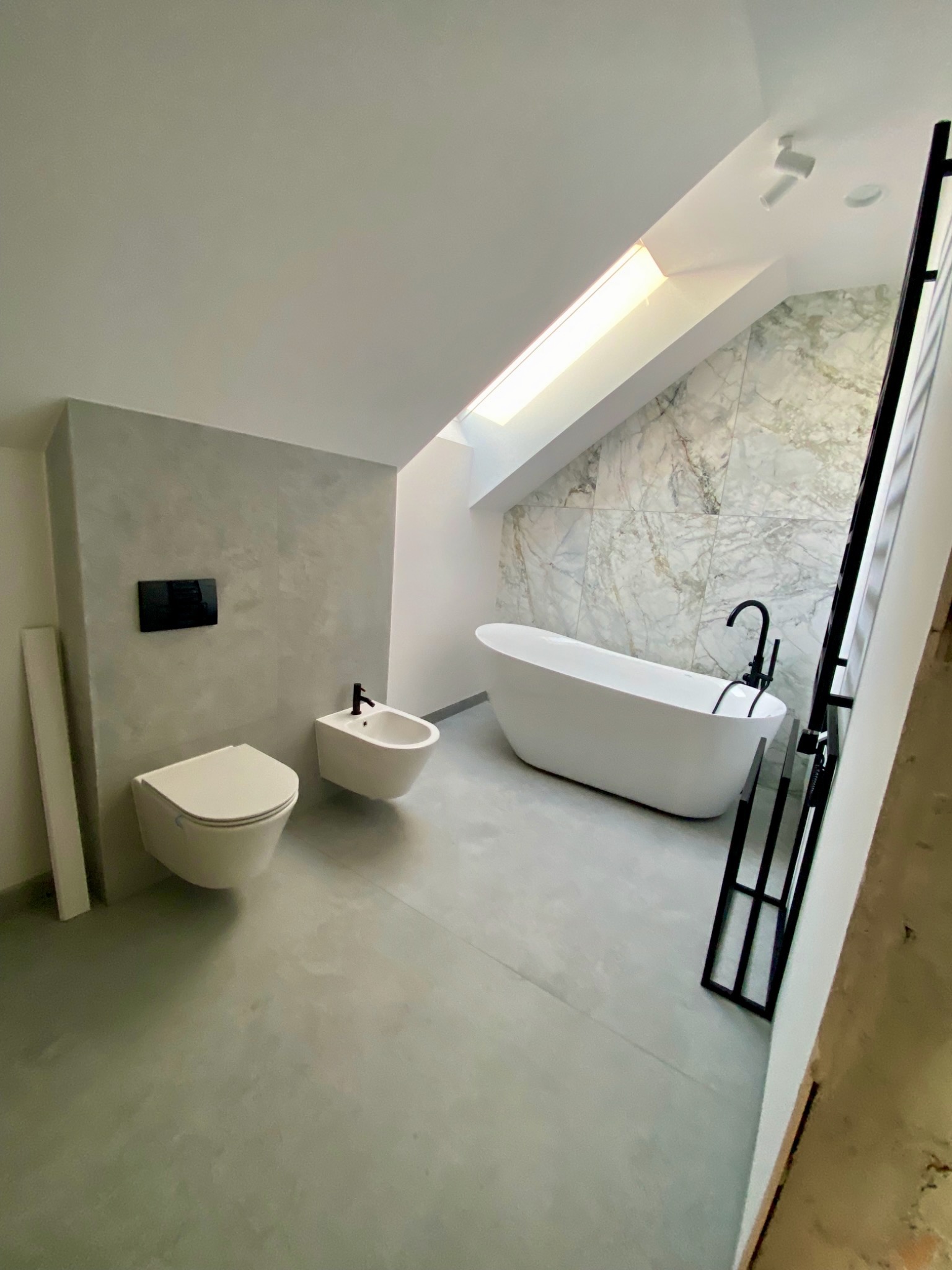 Nowoczesna łazienka na poddaszu z wolnostojącą wanną, bidetem i toaletą w szaro-białej tonacji. Marmurowe płytki na ścianie i okno dachowe doświetlają wnętrze.