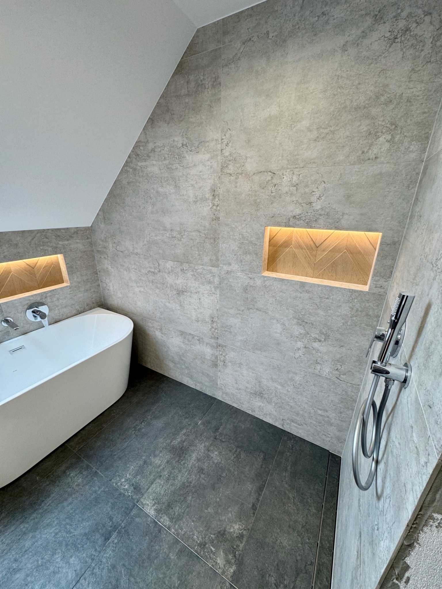 Nowoczesna łazienka w szarościach z wolnostojącą wanną, podświetlanymi niszami z drewnianym wzorem i chromowaną armaturą. Ukośny sufit dodaje unikalnego charakteru.