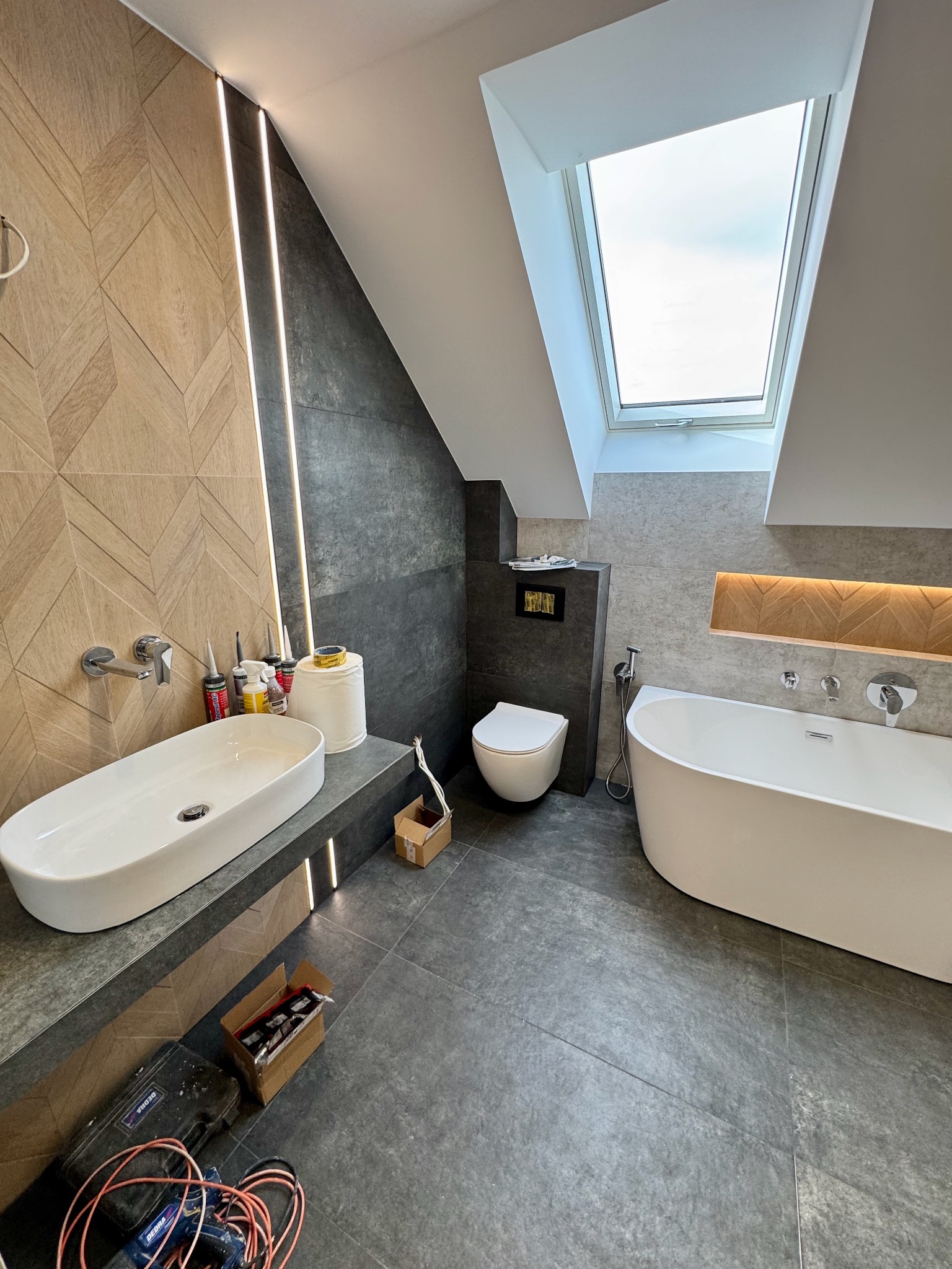 Nowoczesna łazienka na poddaszu z oknem dachowym, wykończona płytkami w odcieniach szarości i beżu. Umywalka nablatowa, wanna i toaleta w minimalistycznym stylu. Widoczne narzędzia pracy.