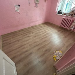 KS BUD - Pusty pokój w Świdnicy z różowymi ścianami o wyraźnej fakturze, drewnianą podłogą i widocznym fragmentem białych drzwi. Na podłodze leżą opakowania po materiałach budowlanych. Widoczny również...