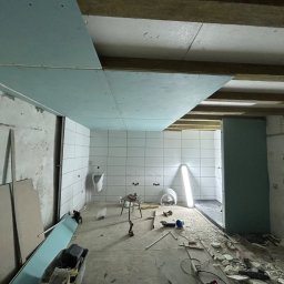 KS BUD - Wnętrze łazienki w trakcie remontu, widoczne płyty kartonowo-gipsowe na suficie, białe płytki na ścianach, zamontowany pisuar, narzędzia i materiały budowlane porozrzucane na podłodze, surowy stan...