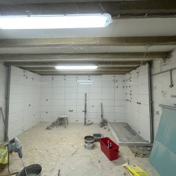 KS BUD - Wnętrze w trakcie remontu, ściany wyłożone białymi płytkami, widoczne narzędzia, wiadro z zaprawą, puszki z piwem w czerwonym pojemniku, surowy strop z widocznymi kablami, pajęczyny w rogach.