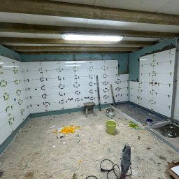 KS BUD - Ściany w trakcie układania białych płytek, z użyciem klipsów poziomujących, widoczne narzędzia, niezagruntowany sufit z drewnianymi belkami i surowa podłoga.