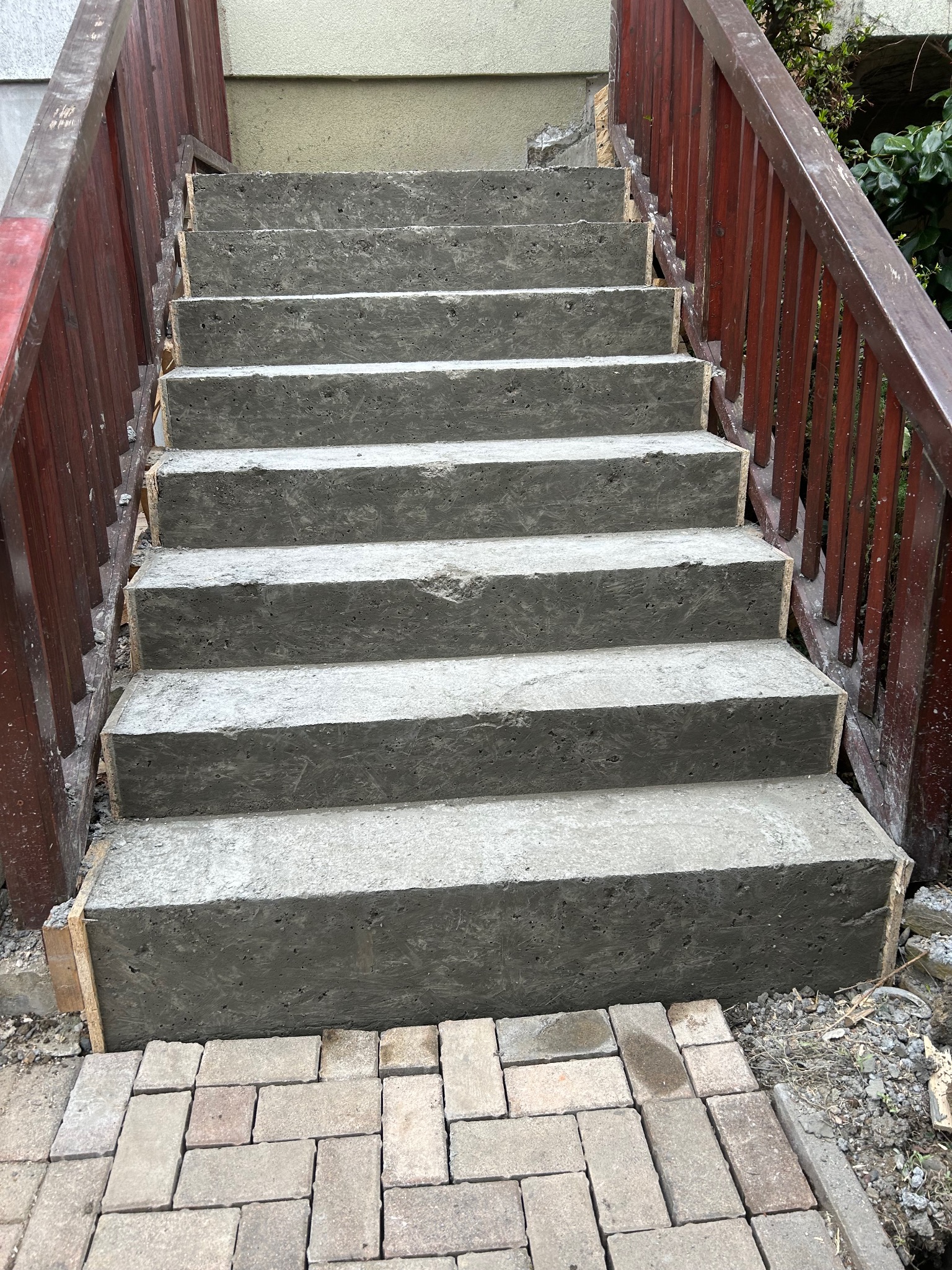 Betonowe schody zewnętrzne w trakcie budowy, z drewnianym szalunkiem i czerwoną drewnianą balustradą, prowadzące w górę, z kostką brukową u podstawy.