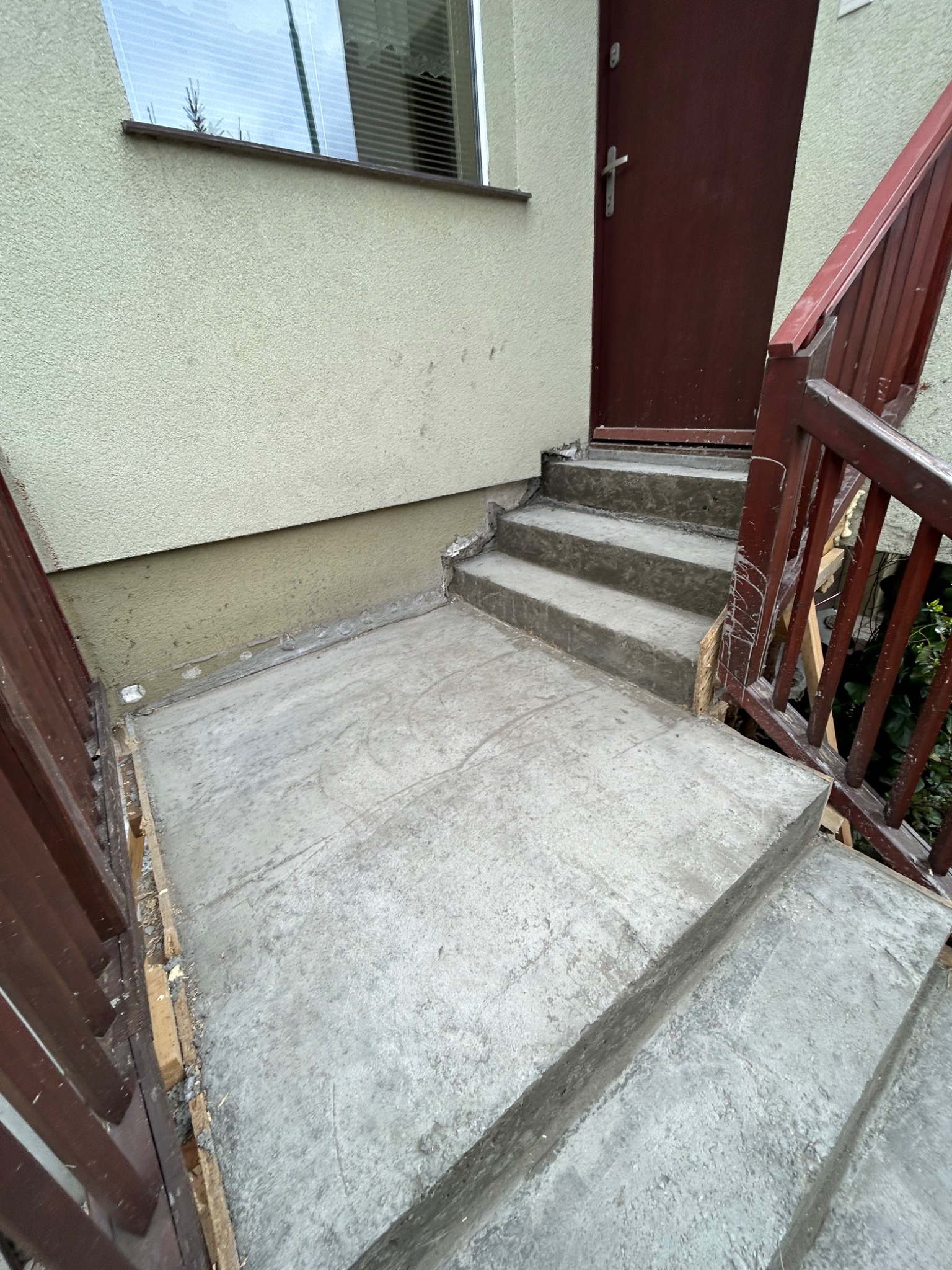 Betonowe schody wejściowe w trakcie remontu, z widocznymi śladami po deskowaniu i przygotowaniach do dalszych prac wykończeniowych, brązowe balustrady po bokach.
