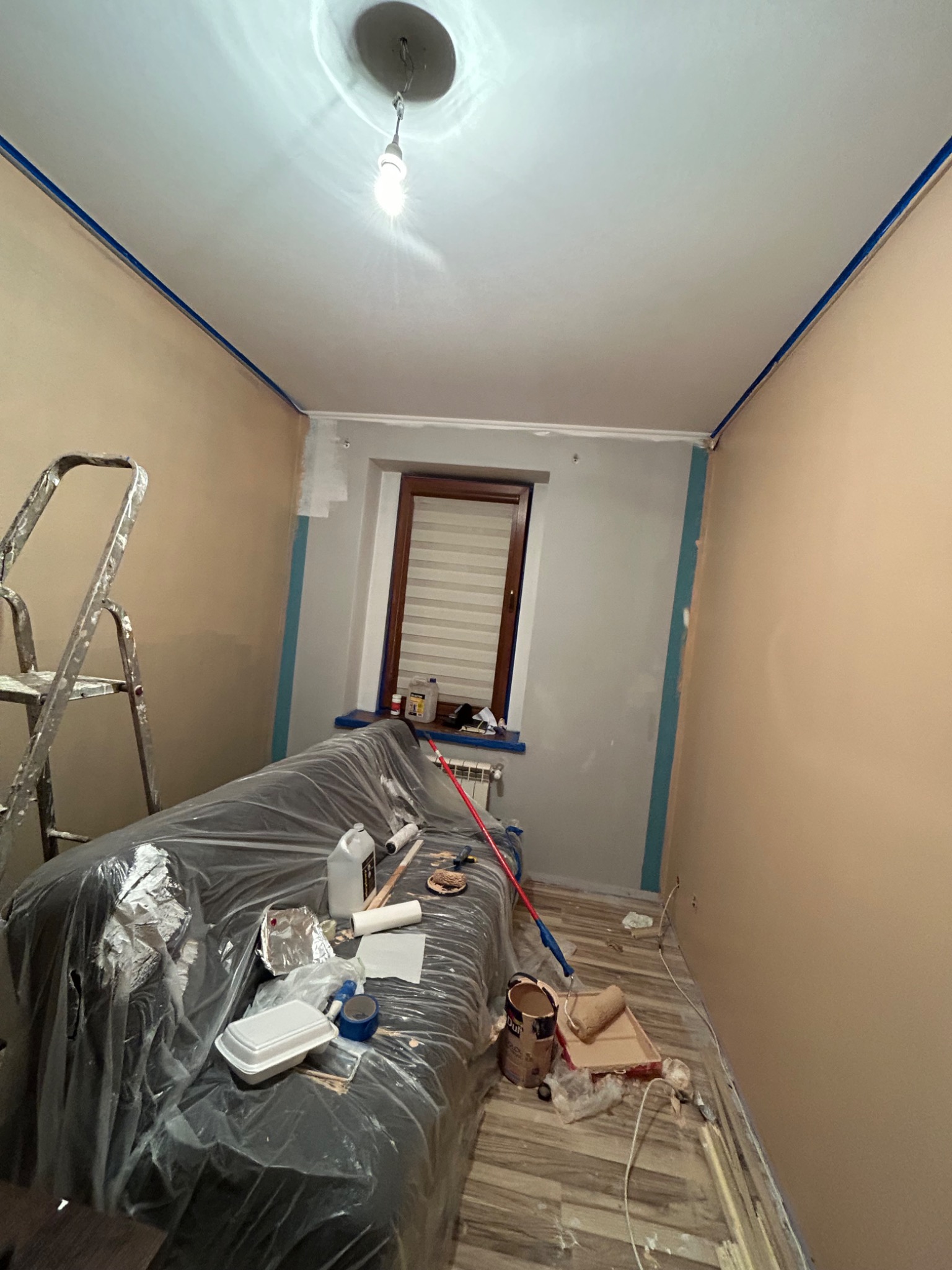 Pokój w trakcie odświeżania: ściany częściowo pomalowane na beżowo, sufit biały z niebieską taśmą malarską przy krawędziach, drabina, zakryta folią kanapa, pędzle i wałki malarskie na podłodze.