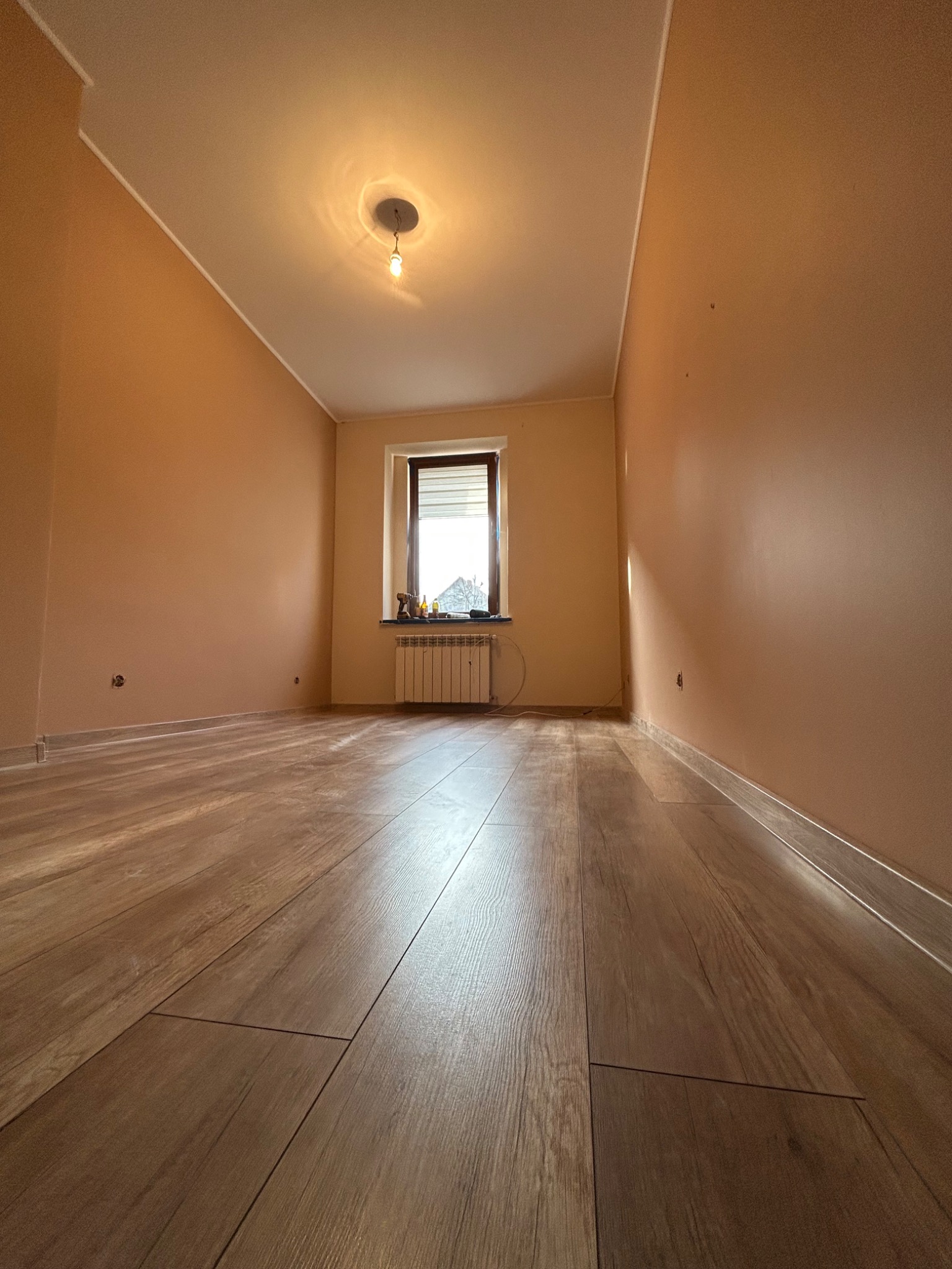 Pusty pokój po remoncie z panelami podłogowymi w odcieniach brązu i ścianami pomalowanymi na ciepły, brzoskwiniowy kolor; widoczne gniazdka elektryczne i grzejnik pod oknem.