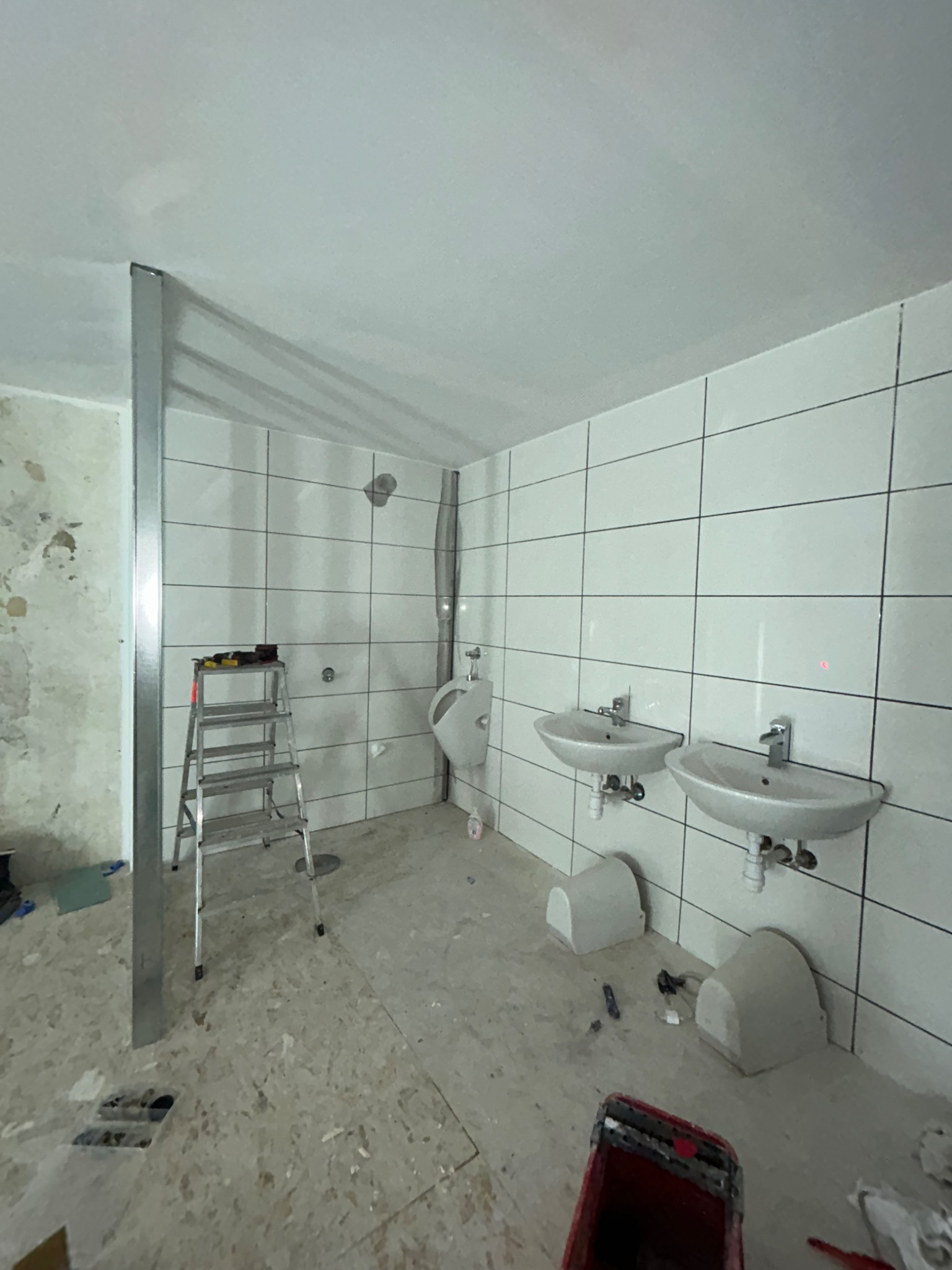 Wnętrze w trakcie remontu: białe płytki na ścianach, dwie umywalki, pisuar, drabina z narzędziami, widoczne ślady prac budowlanych na podłodze i ścianach.