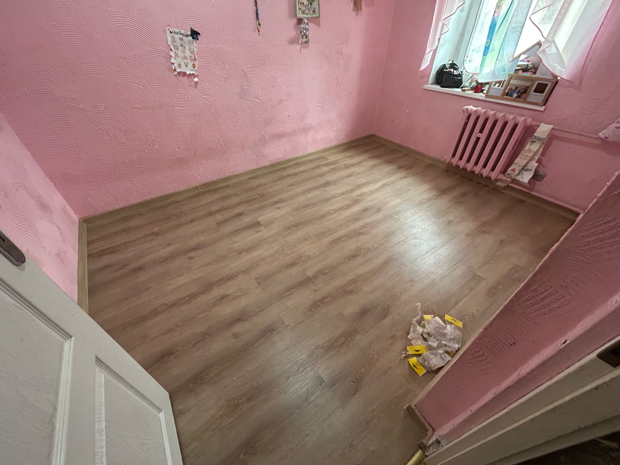 Pusty pokój w Świdnicy z różowymi ścianami o wyraźnej fakturze, drewnianą podłogą i widocznym fragmentem białych drzwi. Na podłodze leżą opakowania po materiałach budowlanych. Widoczny również...