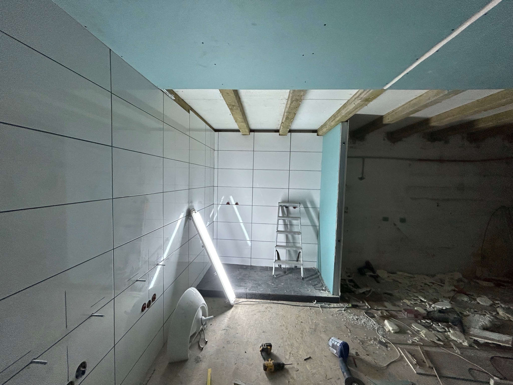 Wnętrze łazienki w trakcie remontu: białe płytki na ścianach, surowy sufit z drewnianymi belkami, prowizoryczne oświetlenie halogenowe, drabina i elementy wyposażenia sanitarnego na podłodze...