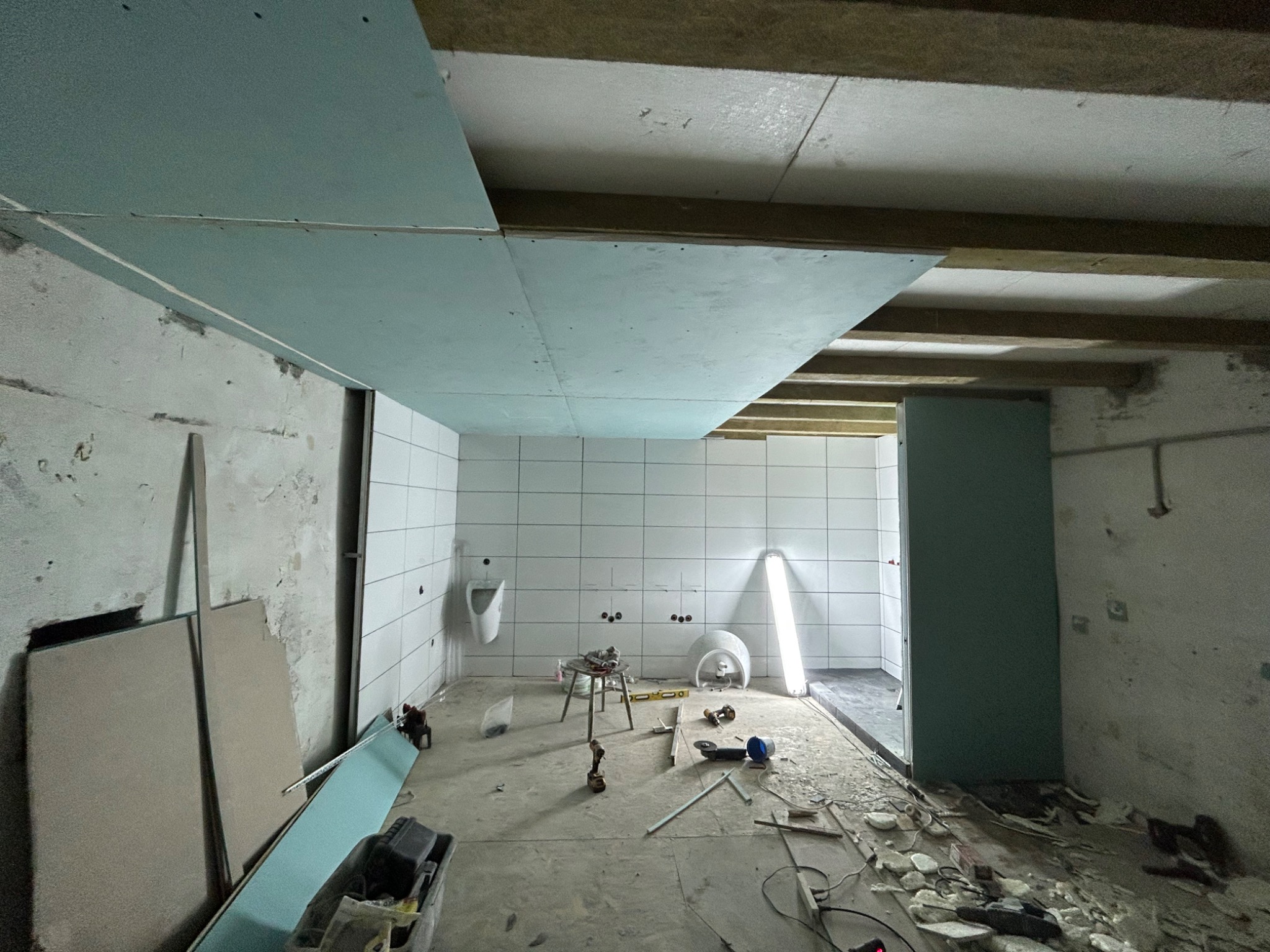 Wnętrze łazienki w trakcie remontu, widoczne płyty kartonowo-gipsowe na suficie, białe płytki na ścianach, zamontowany pisuar, narzędzia i materiały budowlane porozrzucane na podłodze, surowy stan...