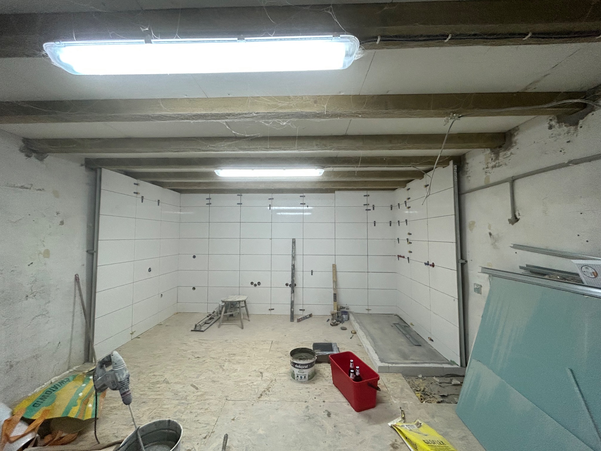 Wnętrze w trakcie remontu, ściany wyłożone białymi płytkami, widoczne narzędzia, wiadro z zaprawą, puszki z piwem w czerwonym pojemniku, surowy strop z widocznymi kablami, pajęczyny w rogach.