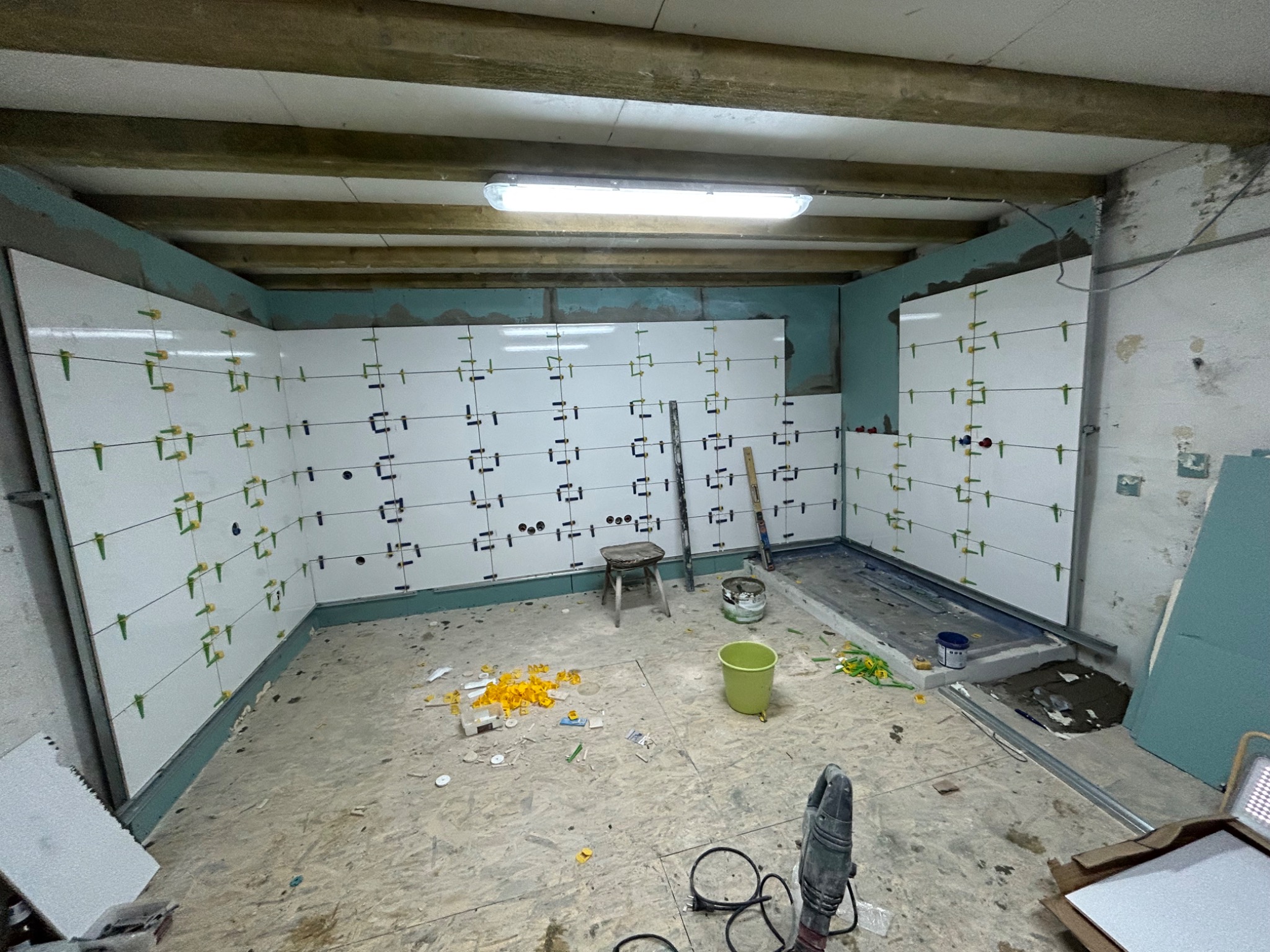 Ściany w trakcie układania białych płytek, z użyciem klipsów poziomujących, widoczne narzędzia, niezagruntowany sufit z drewnianymi belkami i surowa podłoga.