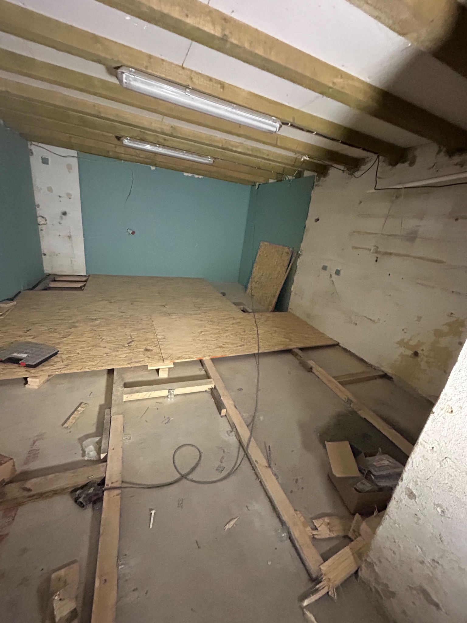 Wnętrze w trakcie remontu: surowy strop z drewnianych belek i lampami, fragment podłogi wyłożony płytami OSB na legarach, odsłonięte ściany z resztkami farby i tynku, narzędzia i materiały...