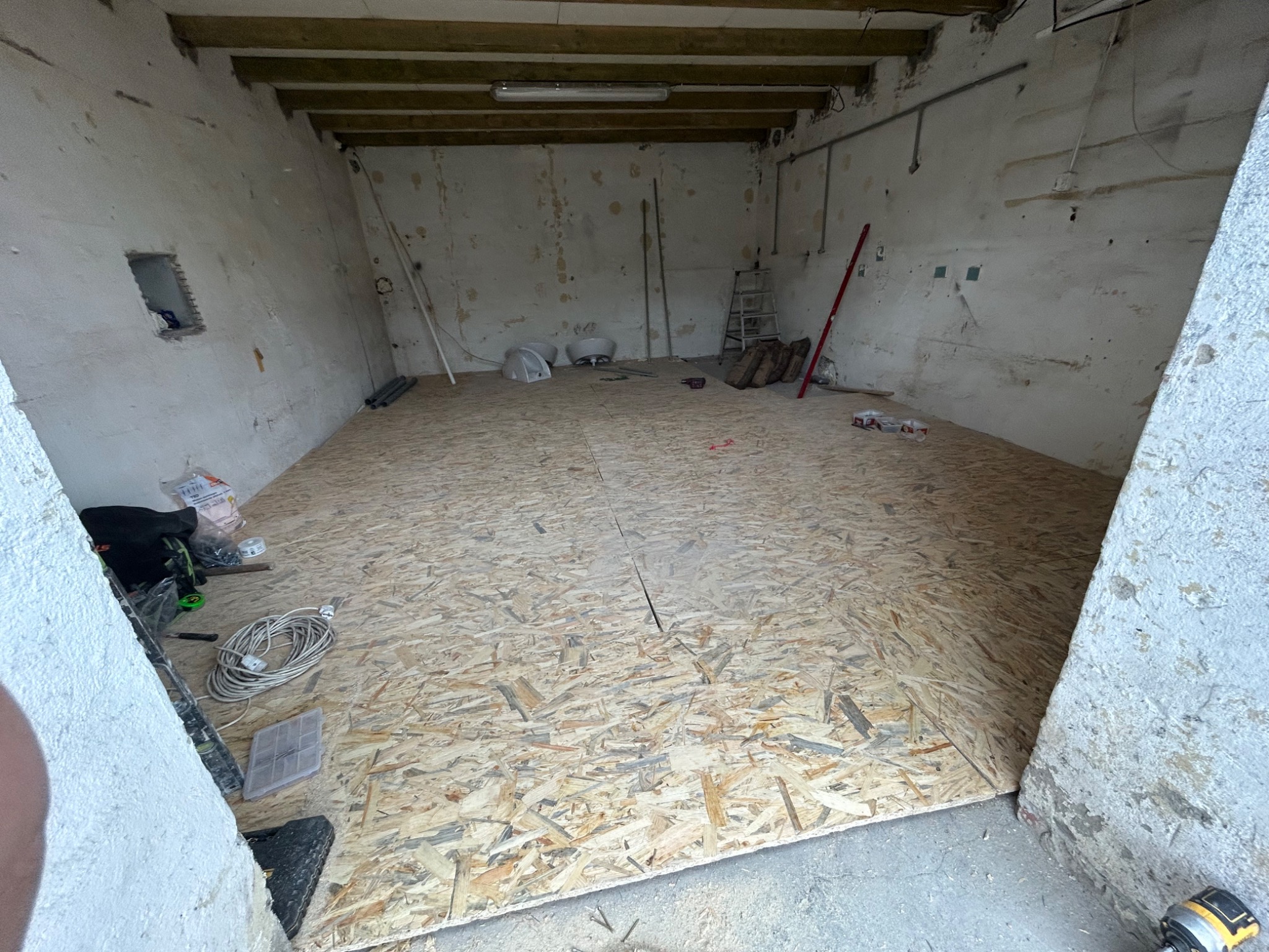 Wnętrze remontowanego pomieszczenia z surowymi ścianami i częściowo ułożoną podłogą z płyt OSB. Widoczne narzędzia, kable i akcesoria budowlane.