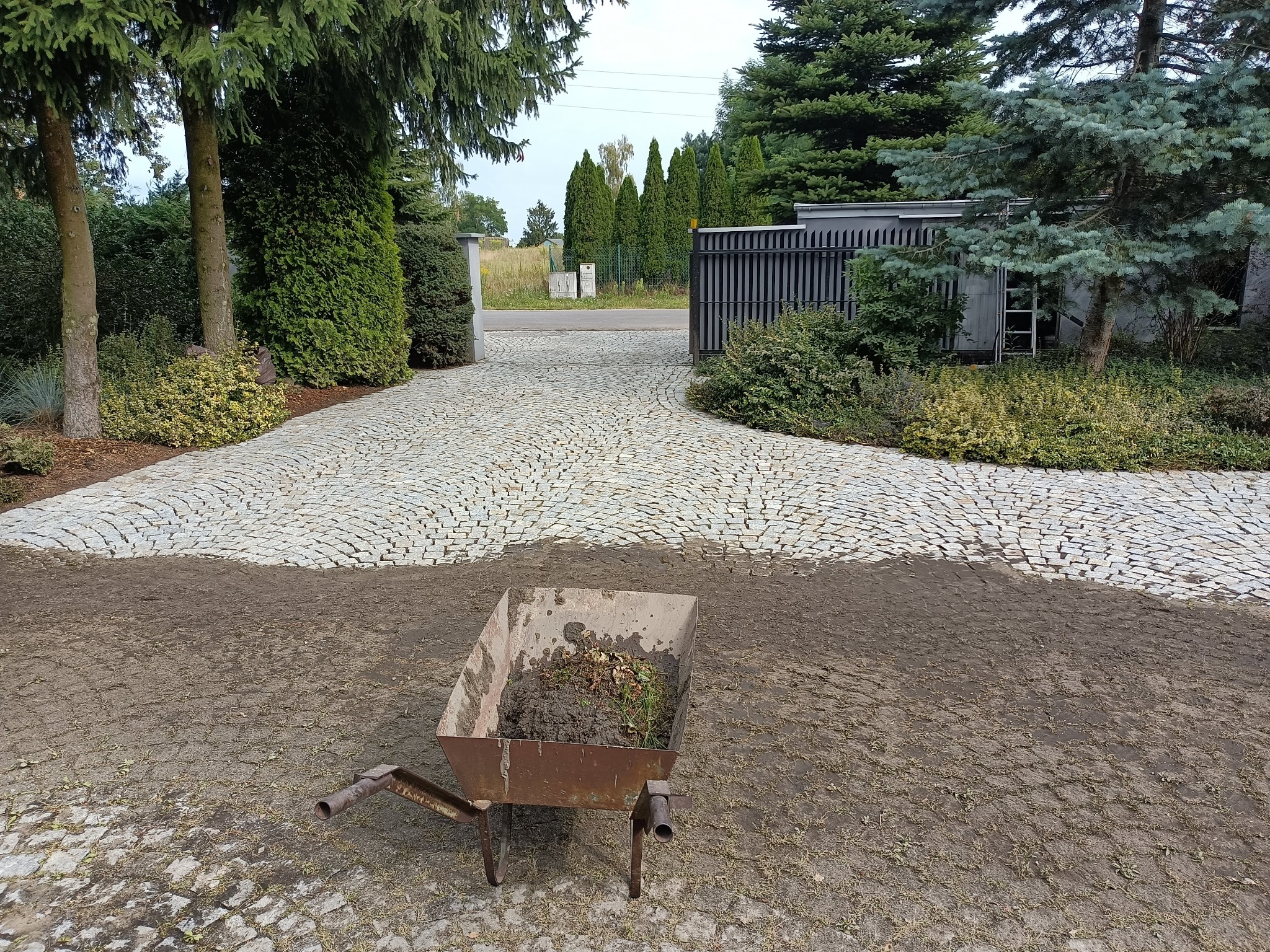 Ułożona kostka brukowa na podjeździe, widoczna taczka z ziemią i resztkami roślin w trakcie prac porządkowych.