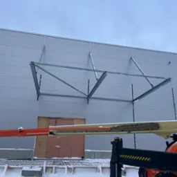 Montaż metalowej konstrukcji wsporczej na ścianie hali z płyt warstwowych, widoczny podnośnik koszowy w zimowej scenerii.