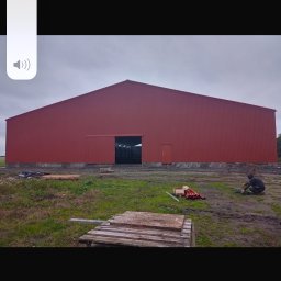 DarStal - Czerwona hala z płyt warstwowych z otwartą bramą przesuwną, widoczna osoba pracująca przy podłożu, palety i rury na trawie.