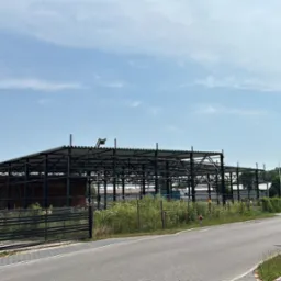 Wznoszona konstrukcja stalowa hali na tle błękitnego nieba, widoczna zza ogrodzenia posesji z domem jednorodzinnym.