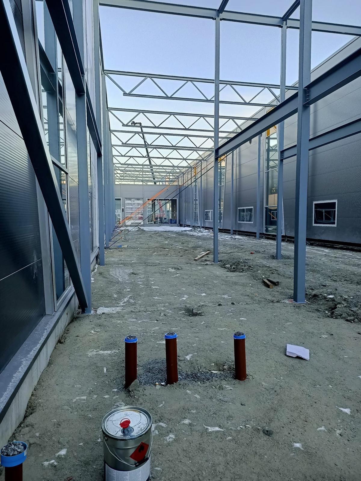 Wnętrze budowanej hali z widoczną stalową konstrukcją dachu, ścianami z paneli, trzema brązowymi rurami z niebieskimi zaślepkami, puszką farby i kartką papieru na ziemi.