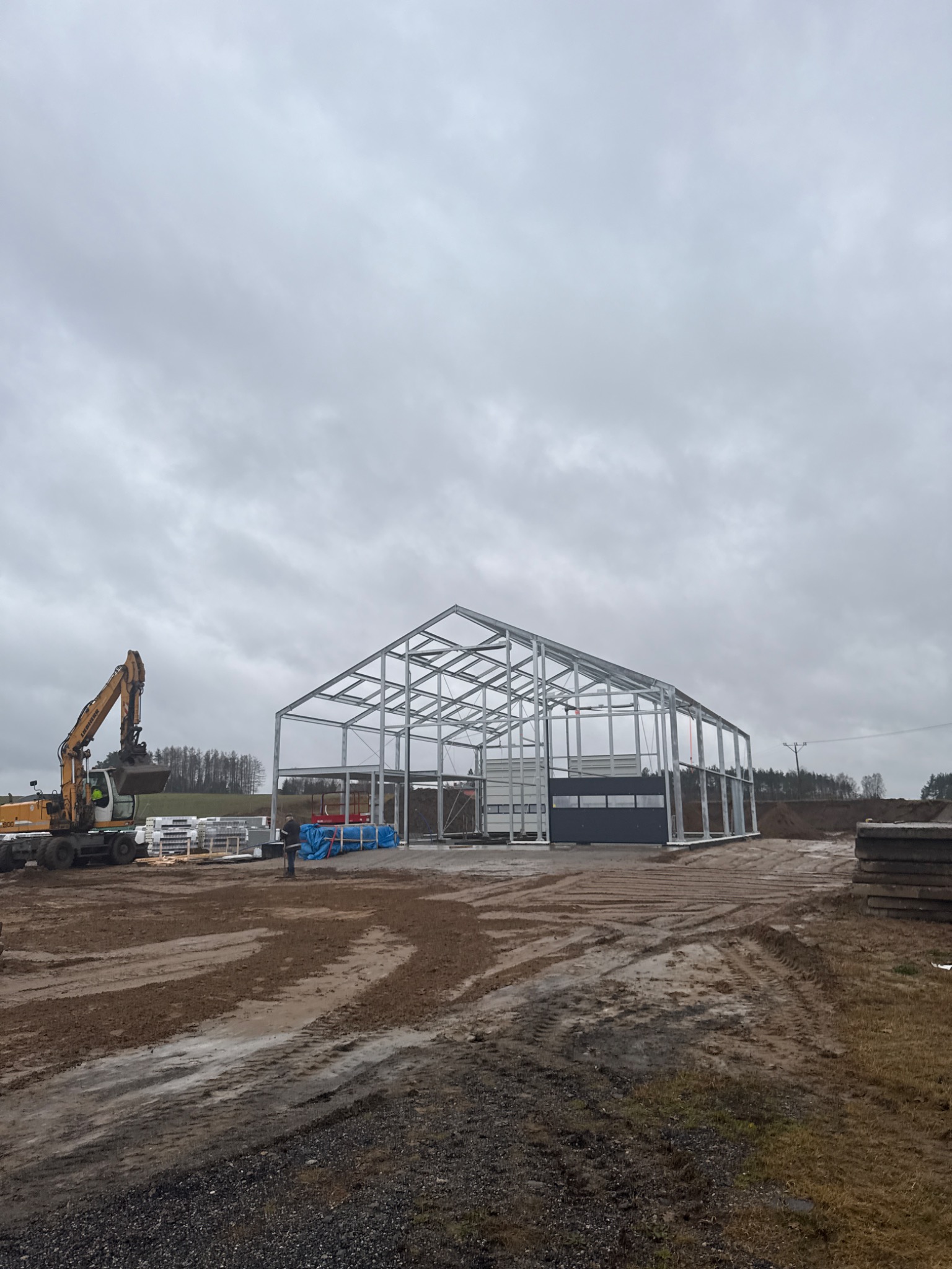 Szkielet stalowej hali w trakcie budowy, widoczny żółty koparka Liebherr 916 i materiały budowlane na błotnistym terenie, pochmurne niebo.