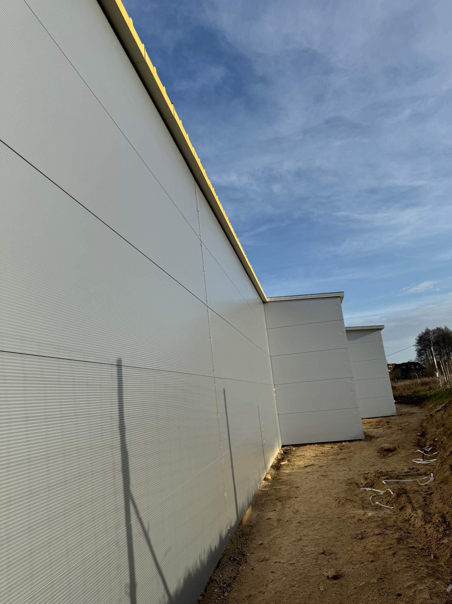 Ściana hali przemysłowej z szarych płyt warstwowych, widok z dołu wzdłuż ściany, na ziemi widoczne ślady budowy.