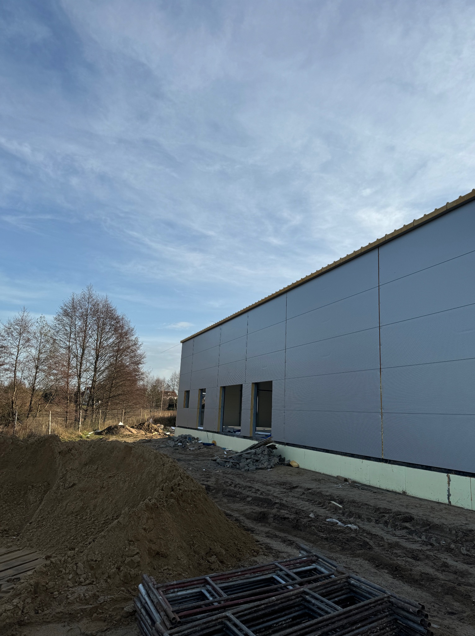 Budynek hali w trakcie budowy, widoczne płyty warstwowe na ścianach, wykop ziemny i metalowe rusztowania na pierwszym planie, błękitne niebo z chmurami w tle.