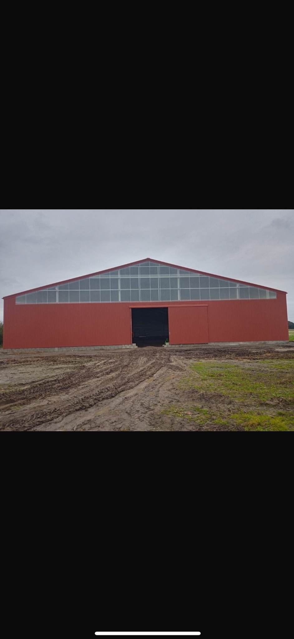 Zewnętrzny widok nowo wybudowanej hali magazynowej z czerwoną elewacją z blachy trapezowej i przeszklonym dachem szczytowym, stojącej na błotnistym terenie.