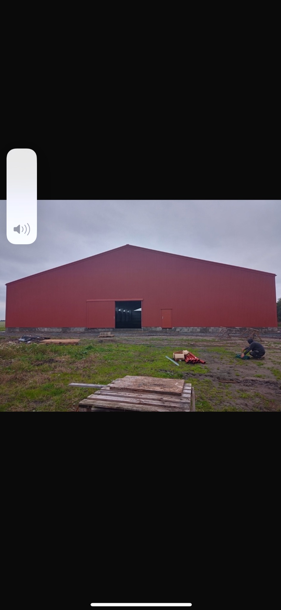 Czerwona hala z płyt warstwowych z otwartą bramą przesuwną, widoczna osoba pracująca przy podłożu, palety i rury na trawie.