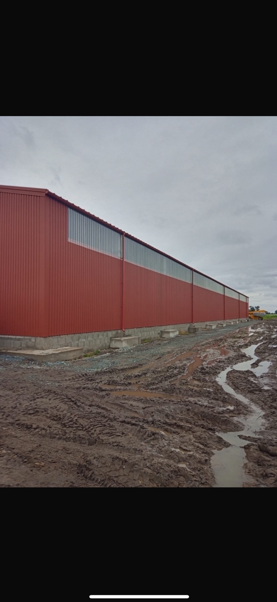 Długa, czerwona hala z blachy trapezowej z pasem okien pod dachem, widoczna z zewnątrz, teren wokół rozjeżdżony i błotnisty, w tle widoczny sprzęt budowlany.
