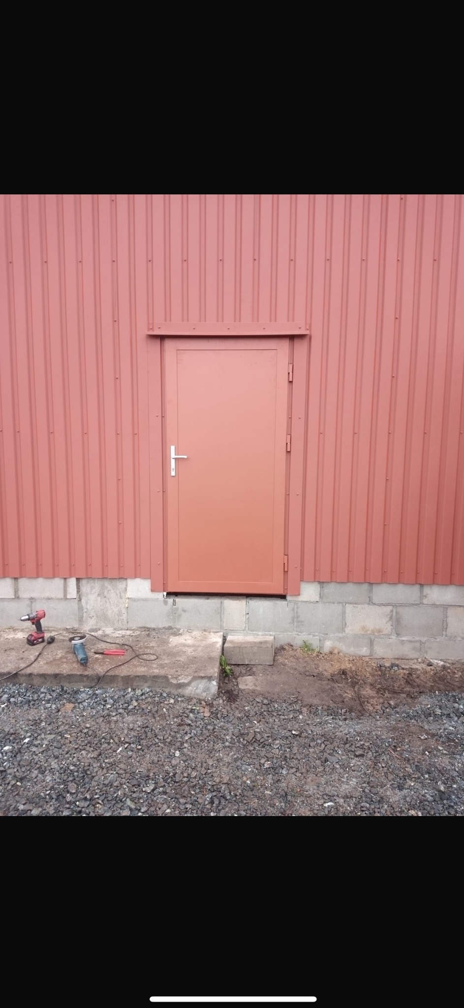 Zamknięte, brązowe drzwi stalowe w budynku z blachy trapezowej, narzędzia budowlane leżą na betonowym podeście.