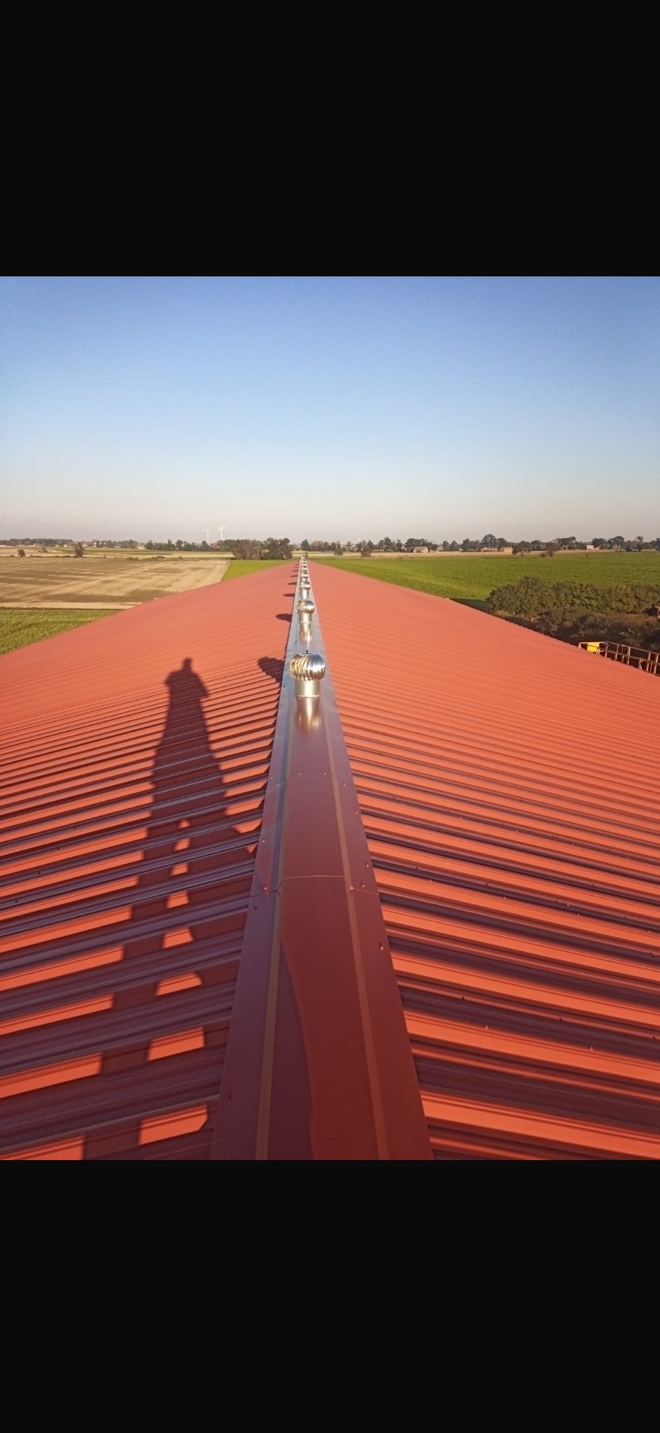 Czerwony dach hali przemysłowej z turbinami wentylacyjnymi w rzędzie, cień osoby na dachu, widok na pola uprawne w oddali.